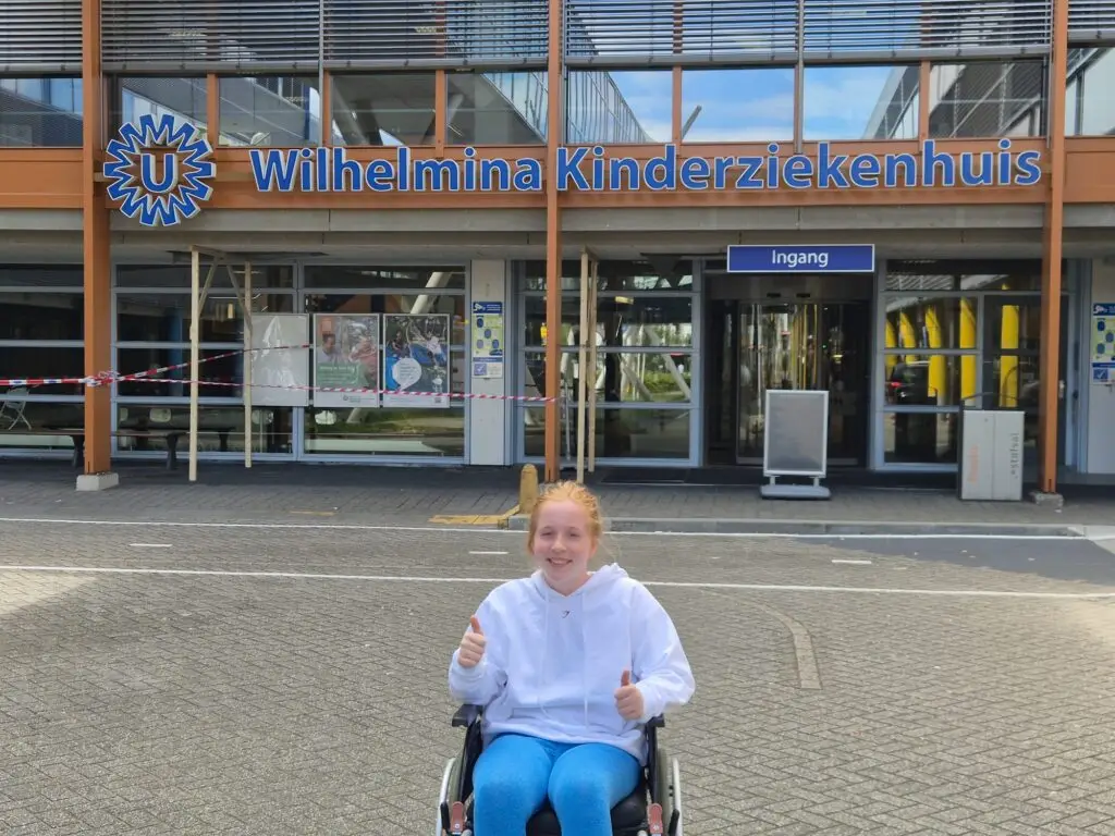  Merel in een rolstoel met duimen omhoog voor de ingang van het Wilhelmina Kinderziekenhuis