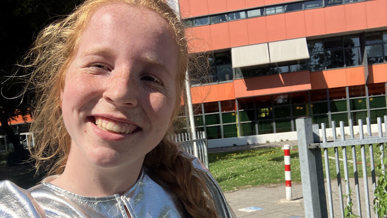 Merel in een zilver shirt, met op de achtergrond een oranje gebouw