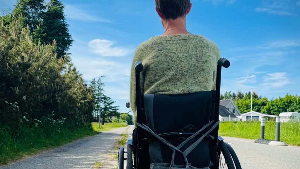 een jongen met kort haar zittend in een rolstoel kijkend naar de wereld, op de achtergond is bos en gras te zien met lucht